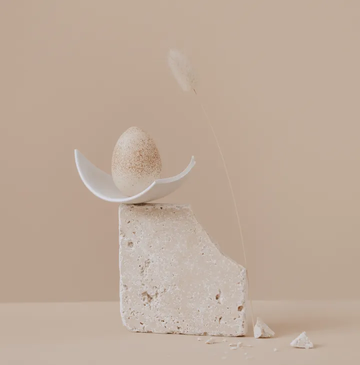 A speckled egg balanced delicately on a half-bowl atop a textured stone block, with a soft beige background and a single dried grass stem.