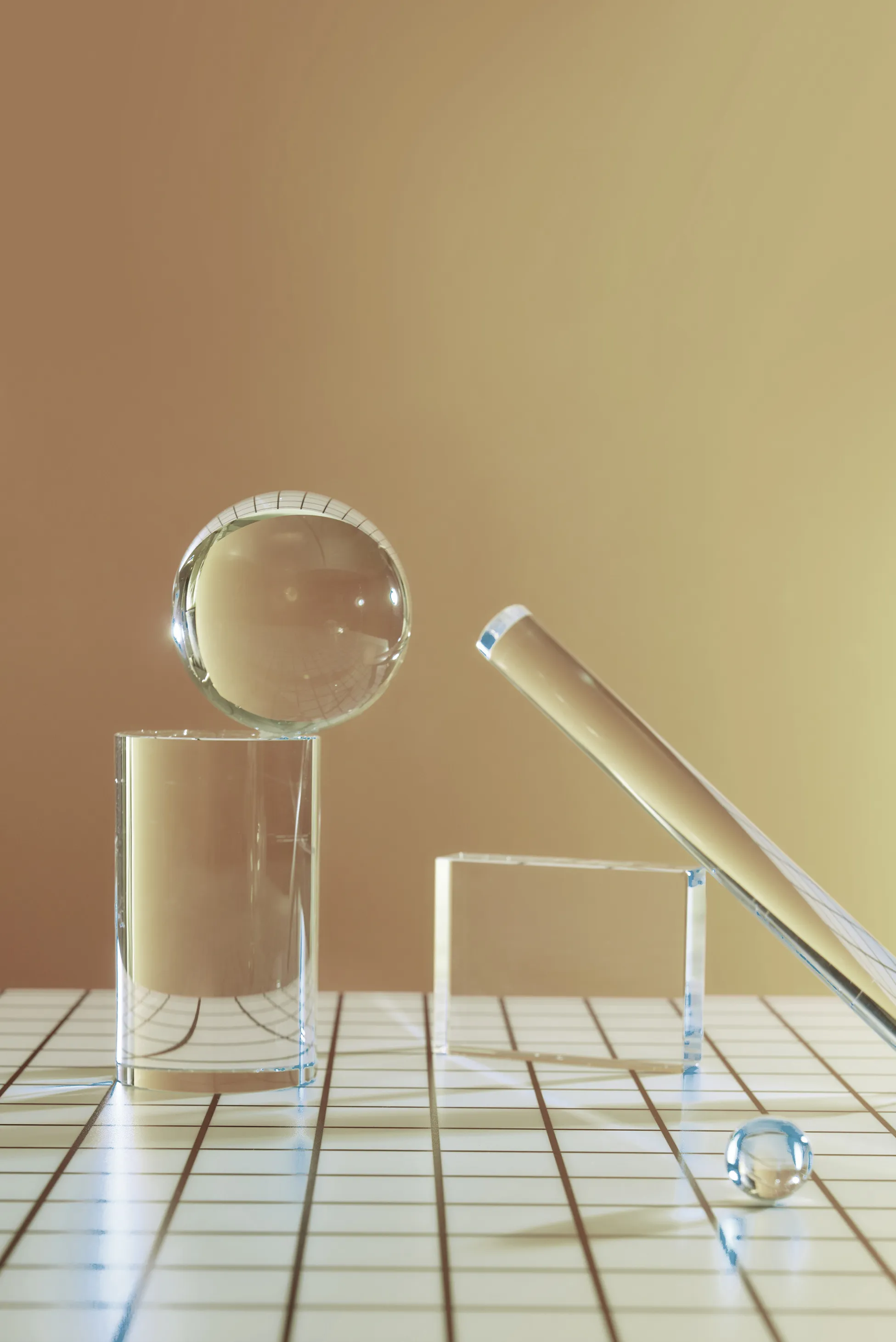A composition of clear glass objects, including a sphere, a cylinder, and a cube on a white grid-patterned surface with a warm beige backdrop.