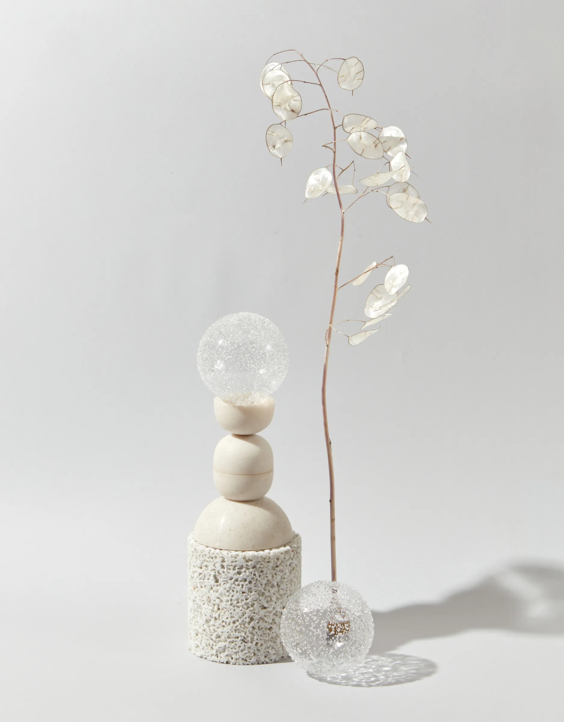 A composition of stacked stones and glittering glass spheres with a delicate dried plant on a shadowed background.